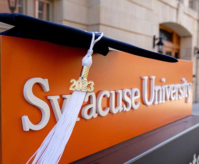 A graduation cap sits on a Syracuse University sign