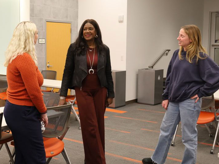 woman on left with blonde hair and wearing orange sweater, center woman has long dark hair wearing a silver necklace over a brown slacks and shirt with a dark short length jacket, on right blonde woman wearing jeans and dark blue sweatshirt they are talking together