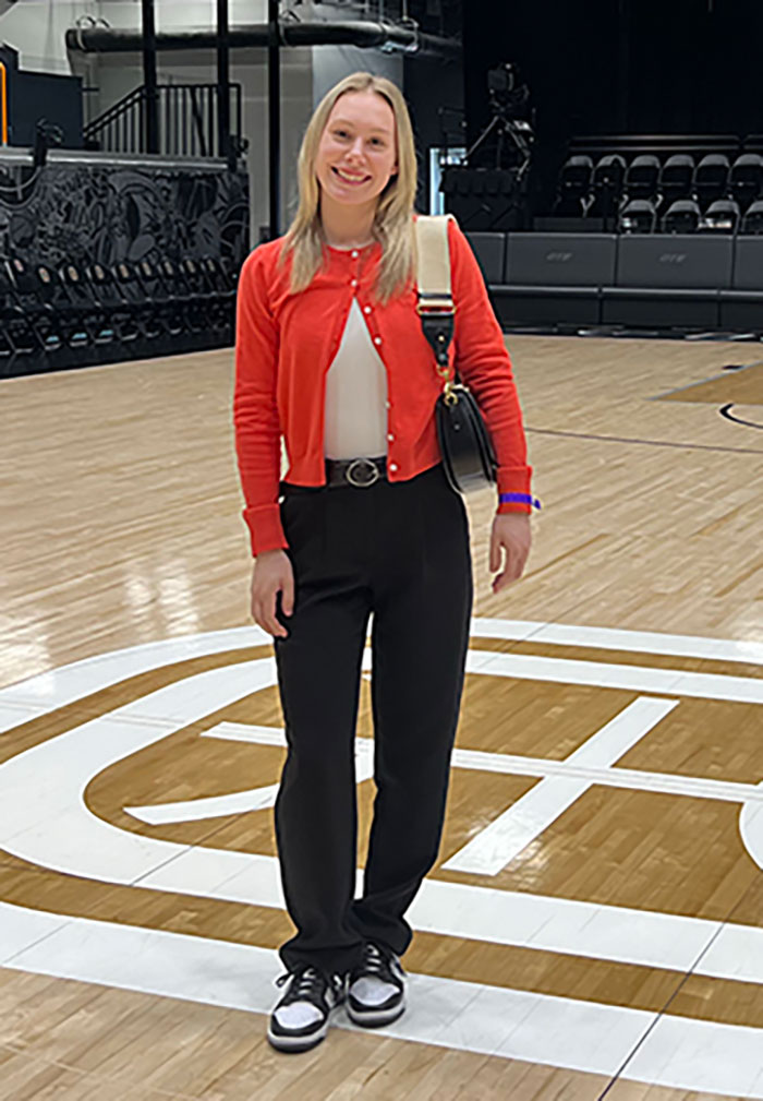 female with long blonde hair wearing dark pants, white shirt and orange sweater standing on basketball court