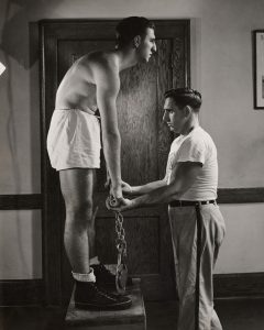 A 1940s photo of a man examining the arm length of a student standing on a stool.