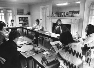 A professor and 6 students are seated around joined tables in a small classroom