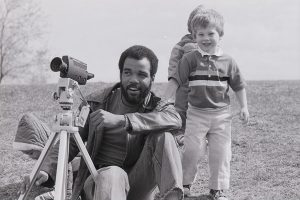 Adult with a 1980s video camera with young children behind him