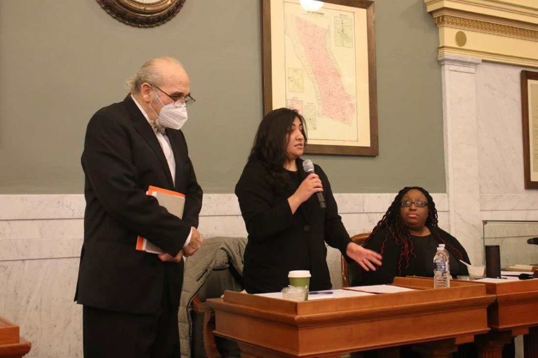 Eric Kingson stands next to a speaker and another person at the front of a courtroom.