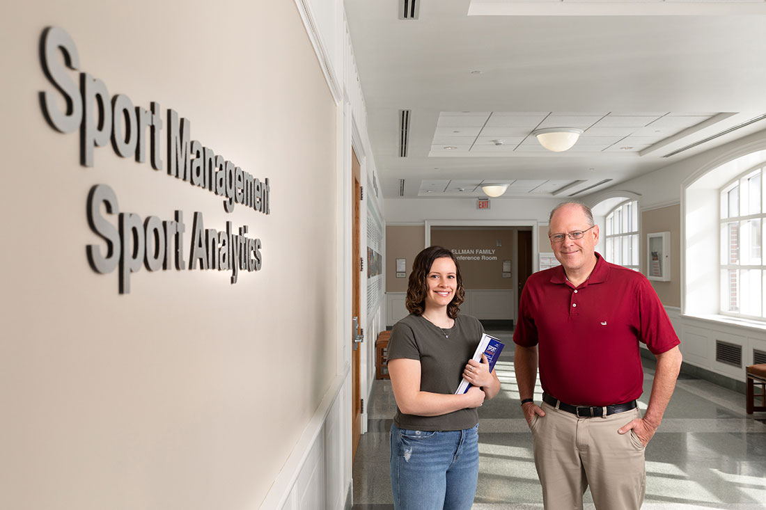 Sport Analytics major and Professor standing in hallway in front of Sport Management adn Sport Analytics sign