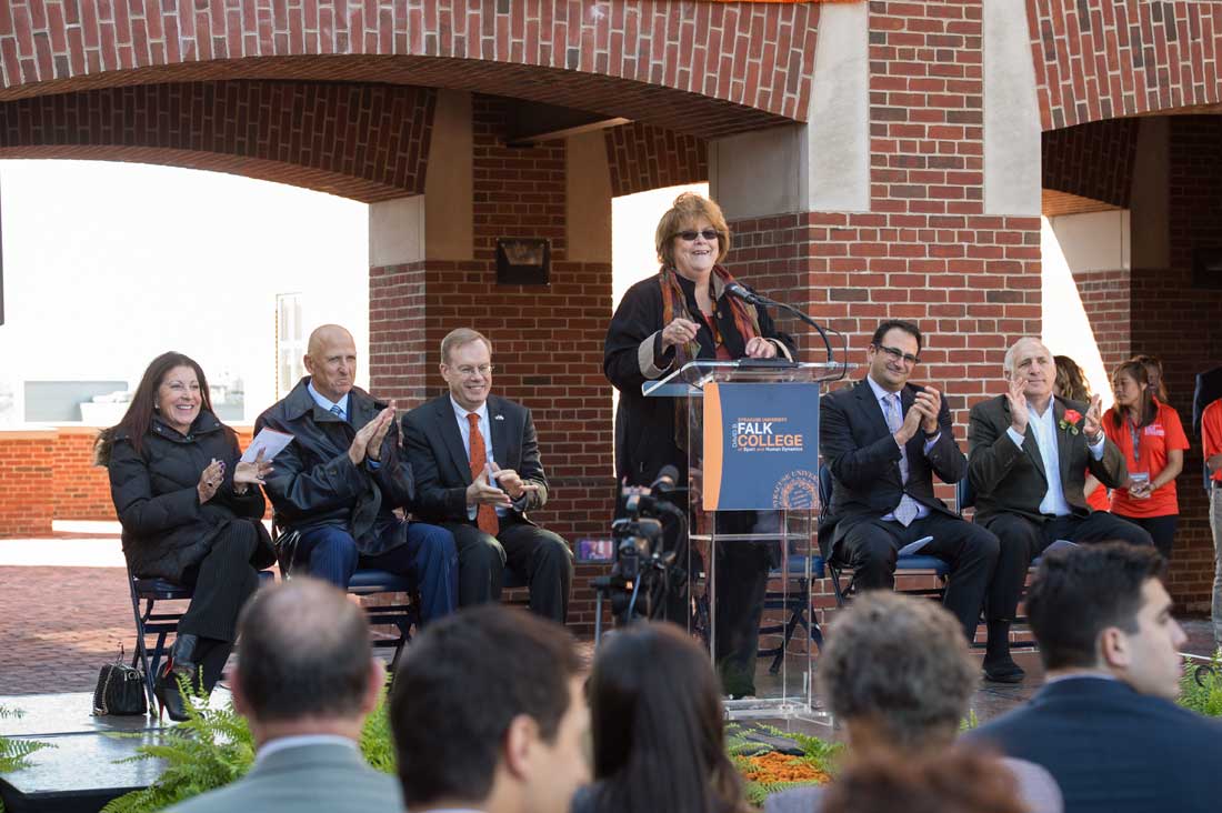 Murphy talks at a podium outside Falk College entrance surrounded by benefactors.