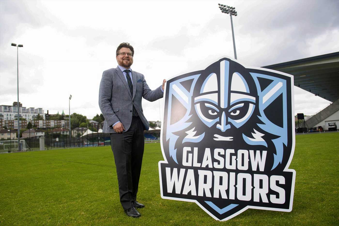 Person stands on rugby pitch and holds a large logo cutout.