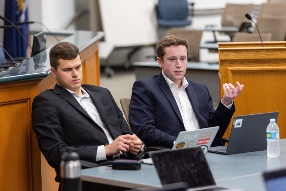 Preston and Jackson are sitting at a table in with laptops discussing with others.