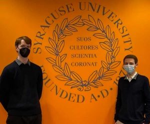 Two young men are posed next to the S.U. seal