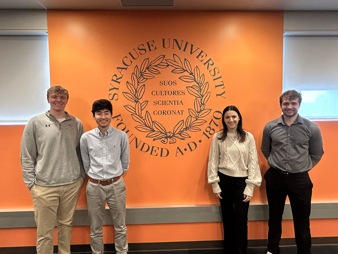 three male members and one female member standing in from of the Syracuse university logo