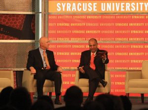 David Falk and Mike Tirico sit on a stage Mike is addressing the crowd