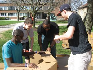 4 male students are putting together a large cardboard box on the S.U. quad