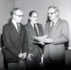 Three men are standing with a paper between them