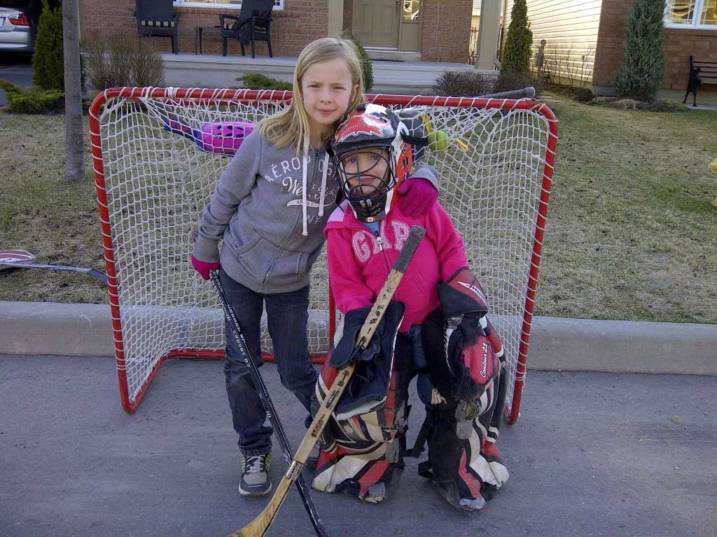 two young girls pose in front of a hockey goal on a street