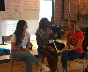 Three Syracuse University students seated in front of brainstorming materials talk with each other at the LIG Playmaker Retreat.