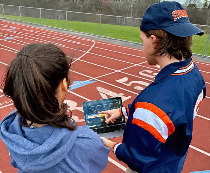 Two students stand on a running track examining a laptop computer screen