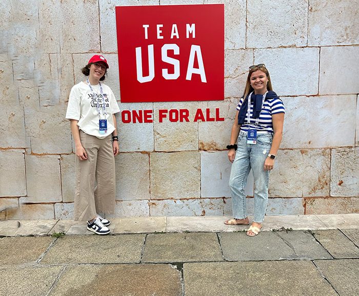 Two women standing in front of a brick wall with red sign that says Team USA one for all