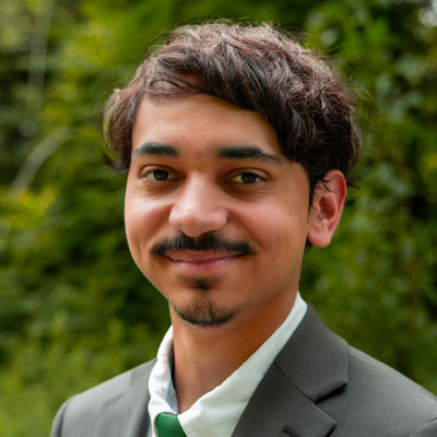 portrait of young man with dark eyes and dark hair standing in front of greenery