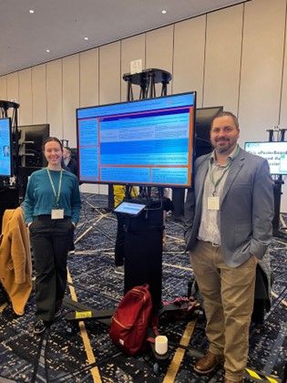 two professors attend conference and are standing on either side of display screen