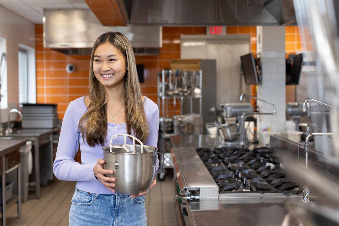 Phoebe Ambrose stands in an industrial kitchen holding a steel mixing bowl.