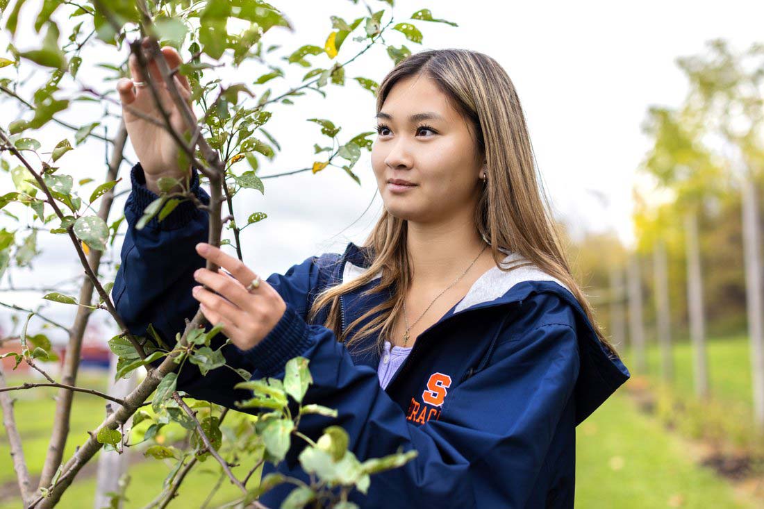 Phoebe Ambrose outside, wearing a coat with the Syracuse University's Block S, holding the branches of a young tree.