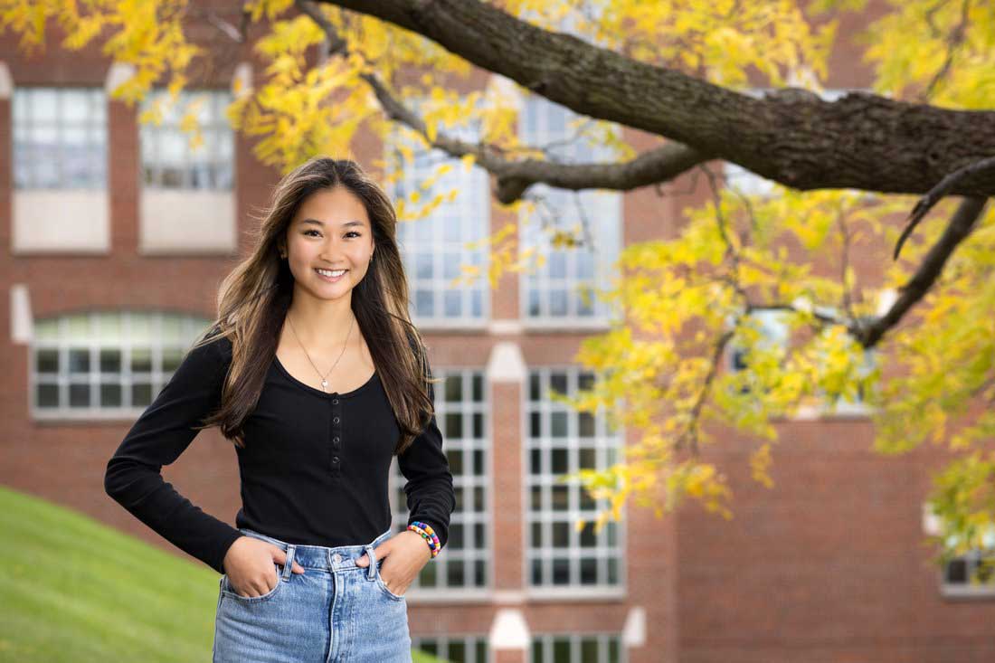 Phoebe Ambrose stands outside under a tree with fall foliage. There's a brick building in the background.