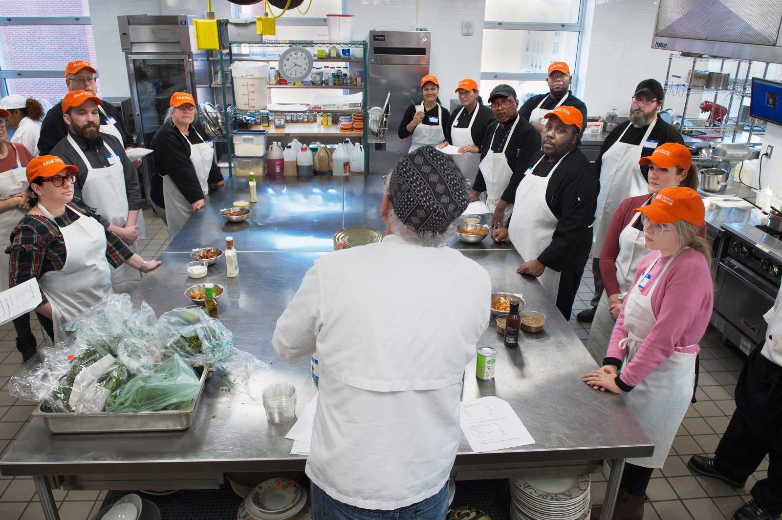 Overhead shot of Chef Bill Collins talks to a roundtable of students in a kitchen